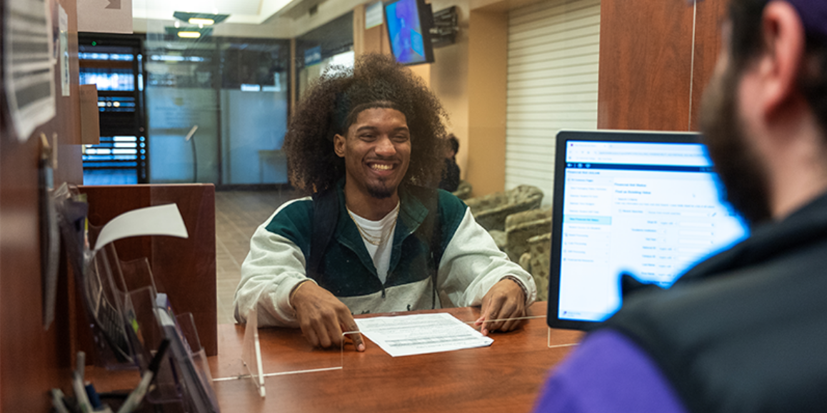 A student at a reception desk talking to someone who has a computer screen open.