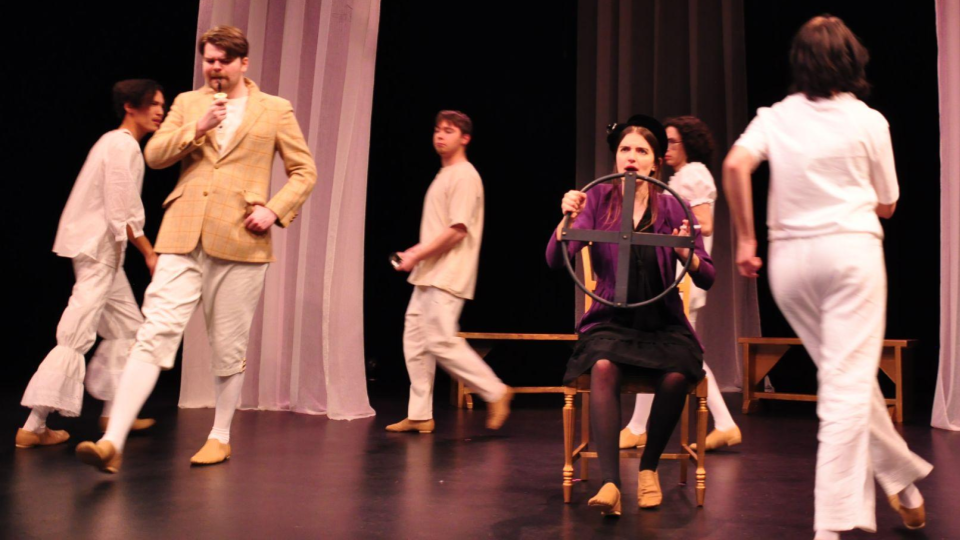 Characters in the musical Alice by Heart. Alfred stands alone watching Alice interact with the colorful caterpillar characters.