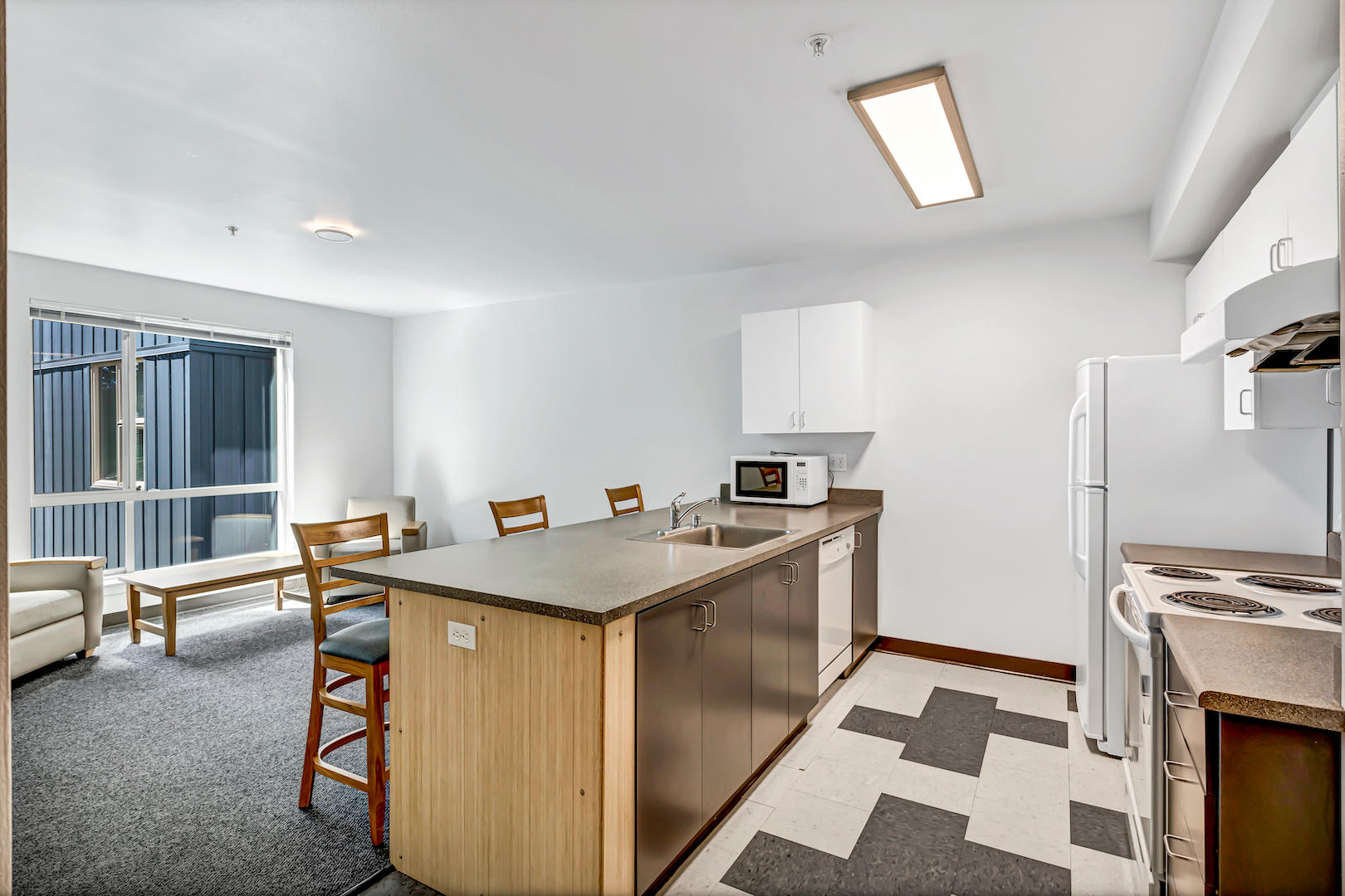 Open-concept kitchen with island seating, white appliances, and adjacent living area with chairs and large window.