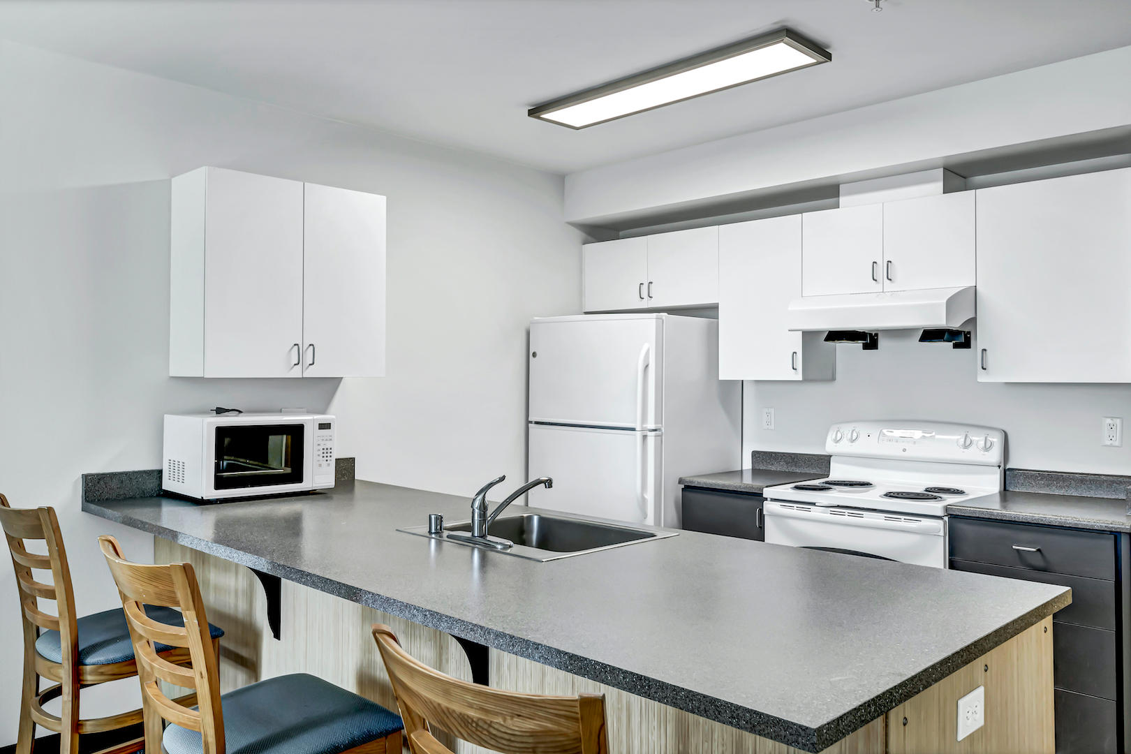 Compact kitchen with white cabinets, electric stove, refrigerator, microwave, and gray countertop.